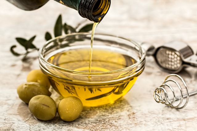 Olivový olej sa leje do misky, vedľa ktorej ležia zelené olivy